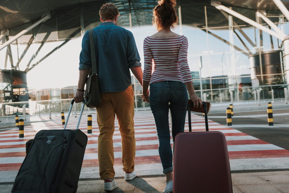 A couple holding hands, pulling suitcases toward an airport terminal