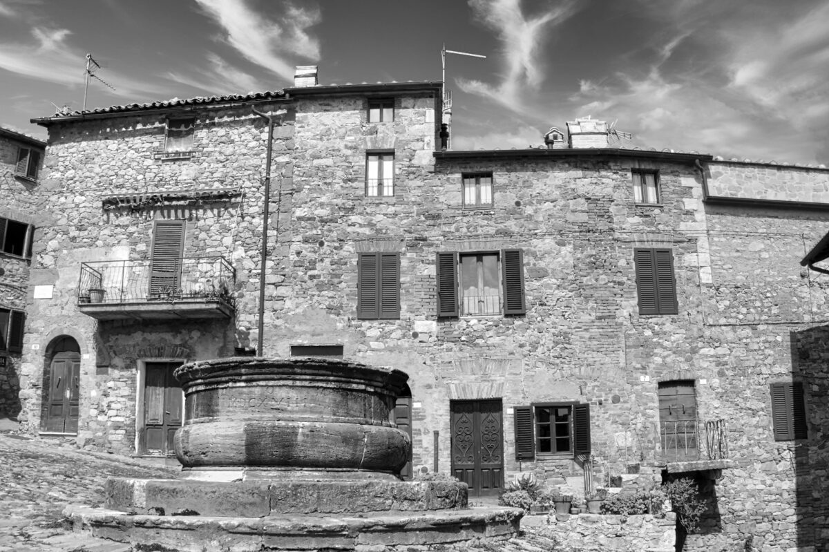 Black-and-white photo of old stone houses around a stone well in an Italian village square